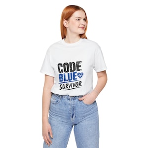 May include: White t-shirt with the text "CODE BLUE SURVIVOR" in black and blue. The design includes a blue heart symbol. The shirt is worn by a person in blue jeans.