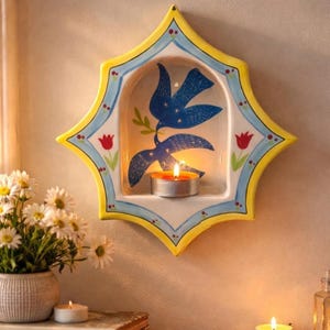 Puede incluir: Aplique de pared de cerámica con forma de estrella, con un diseño de paloma azul que sostiene una rama de olivo. El aplique tiene un borde amarillo, detalles azules y un pequeño portavelas. Una vela encendida está dentro.
