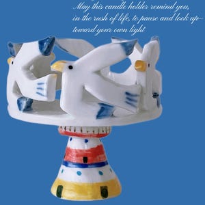 May include: A ceramic candle holder with a lighthouse base and a ring of seagulls. The seagulls are white with blue and yellow details. The lighthouse base is painted with red, white, blue, and yellow stripes. Text reads "Seagulls & Lighthouse".
