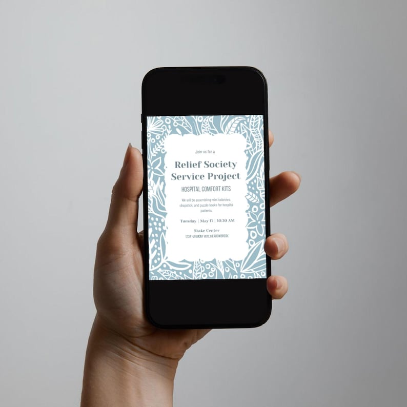 May include: A black smartphone shows a light blue and white invitation for a Relief Society Service Project. The invitation includes text about Hospital Comfort Kits and event details. The phone is held in a hand against a neutral background.