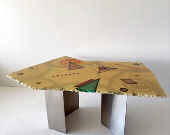 May include: A yellow coffee table with a geometric design painted on the top. The table has a unique, irregular shape and is supported by four silver metal legs.