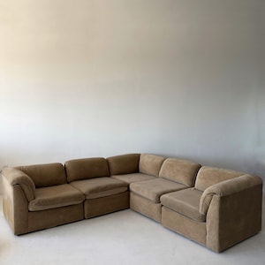 May include: A large, modular sectional sofa in a neutral tan color. The sofa is composed of multiple square and rectangular sections, creating a corner configuration. The upholstery appears to be a soft, suede-like fabric.