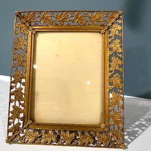 May include: A rectangular gold-toned metal picture frame featuring an intricate openwork border of embossed leaves and small berries. The frame surrounds a plain, off-white rectangular insert and has a textured, metallic finish throughout.
