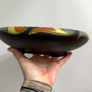 May include: A shallow, circular ceramic bowl with a matte black exterior finish. The rim features a decorative band of stylized, leaf-like shapes in shades of orange, yellow, and teal, applied with a visible, raised texture around the circumference.
