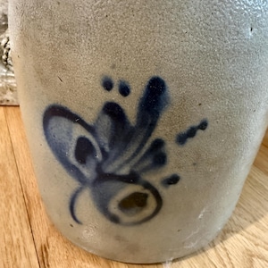 May include: A stoneware jug with a light gray, textured surface. A dark blue floral design is painted on the side. The jug sits on a wooden surface. This antique piece is a decorative item.