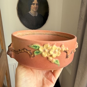May include: A terracotta-colored ceramic bowl with a floral design. The bowl features raised yellow flowers and green leaves on brown branches. The bowl is held in a hand, with a portrait in the background.