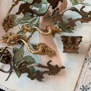 May include: A collection of antique brass hardware, including ornate drawer pulls and a small decorative column. The hardware features intricate floral and scroll designs, with a warm, aged patina. The pieces are arranged on a vintage floral print.