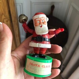 May include: A red and white Santa Claus puppet with a beard and a bell in his hand. The puppet is on a green base with the text "Santa Claus Push Button to Make Him Dance Push Button Underneath Made in Taiwan" printed on it.