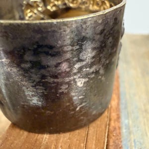 Puede incluir: Primer plano de una taza de metal plateado deslustrado con un borde dorado decorativo. La taza tiene una superficie texturizada y se asienta sobre una superficie de madera. El borde presenta un patr&oacute;n ornamental y repetitivo.
