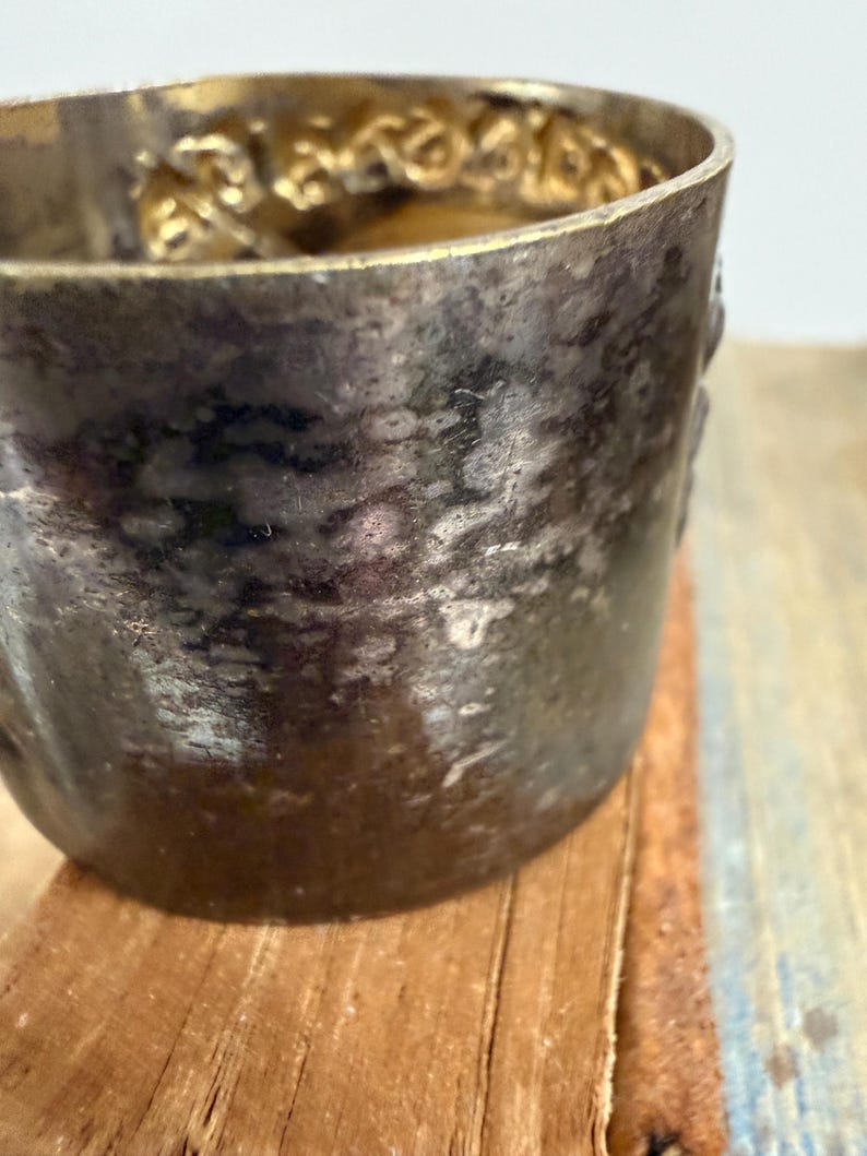 Puede incluir: Primer plano de una taza de metal plateado deslustrado con un borde dorado decorativo. La taza tiene una superficie texturizada y se asienta sobre una superficie de madera. El borde presenta un patr&oacute;n ornamental y repetitivo.