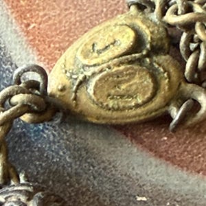 May include: Close-up of a vintage brass pendant with an ornate design. The pendant is attached to a chain and features a detailed, textured surface with swirling patterns. The metal has an aged, antique appearance.