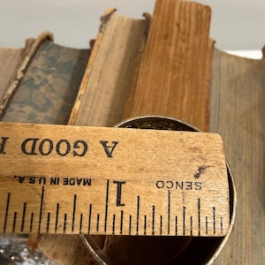 Puede incluir: Un objeto met&aacute;lico, posiblemente una taza o recipiente, se mide con una regla de madera. La regla, marcada en pulgadas, dice "A GOOD" y "MADE IN U.S.A." El objeto mide aproximadamente 2,5 cm de di&aacute;metro. El fondo incluye p&aacute;ginas de libros antiguos.