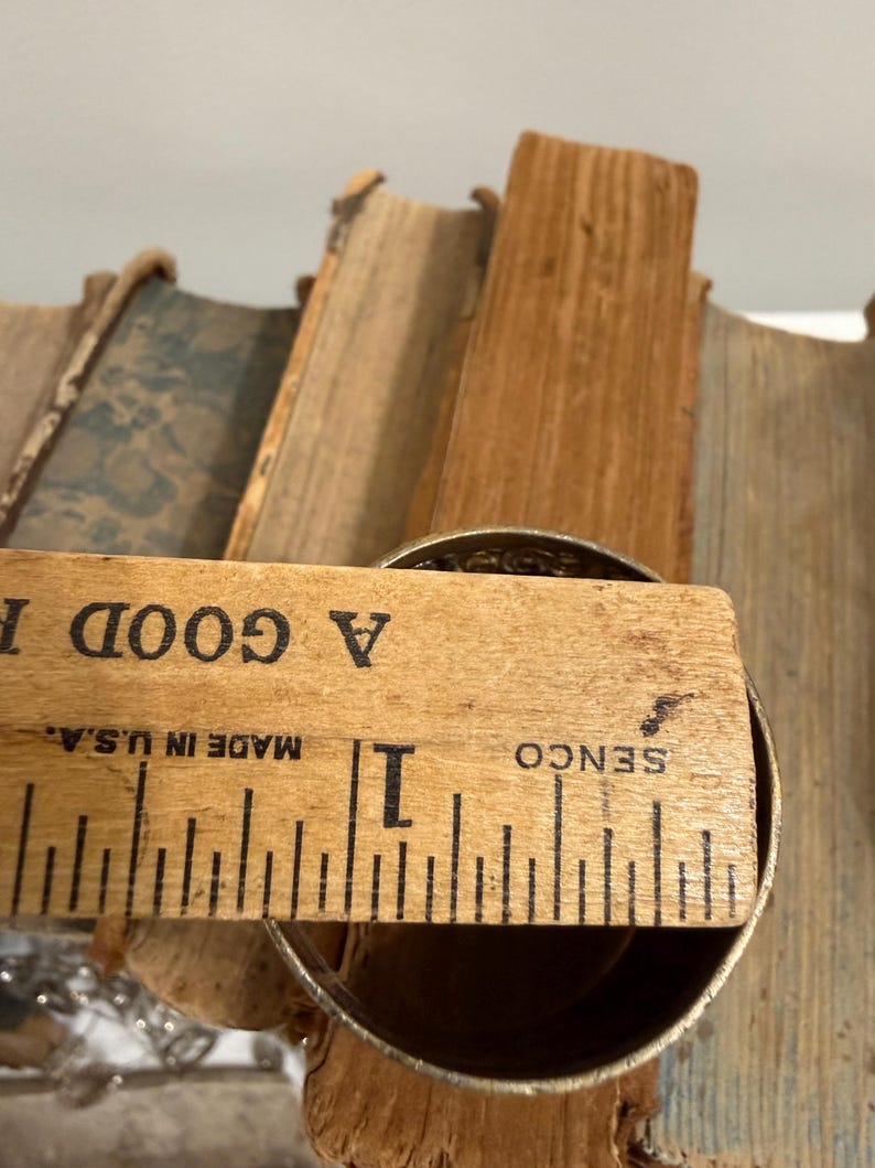 Puede incluir: Un objeto met&aacute;lico, posiblemente una taza o recipiente, se mide con una regla de madera. La regla, marcada en pulgadas, dice "A GOOD" y "MADE IN U.S.A." El objeto mide aproximadamente 2,5 cm de di&aacute;metro. El fondo incluye p&aacute;ginas de libros antiguos.