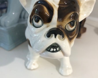 May include: A white and brown ceramic figurine of a French bulldog with a grumpy expression. The figurine has black eyes and a black nose.