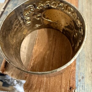 Puede incluir: Un servilletero de metal plateado con un dise&ntilde;o floral en relieve ornamentado. El anillo tiene una banda ancha y una apariencia ligeramente envejecida. El interior tiene un &oacute;valo de color dorado. El servilletero descansa sobre una pila de p&aacute;ginas de libros viejos y desgastados.
