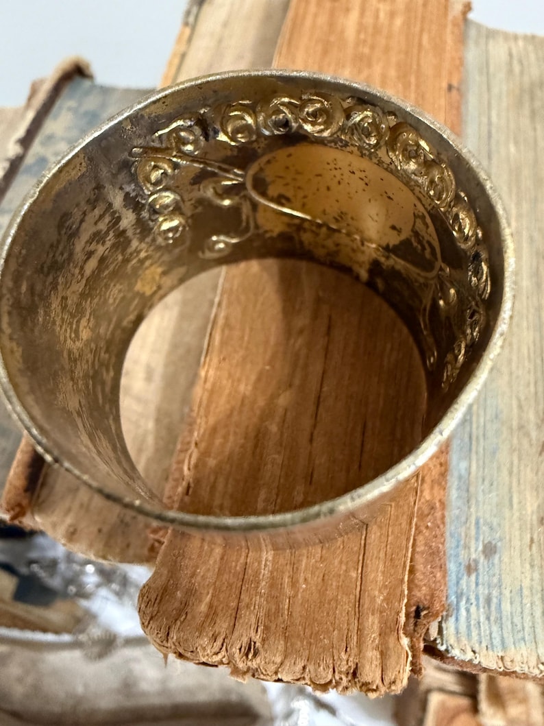 Puede incluir: Un servilletero de metal plateado con un dise&ntilde;o floral en relieve ornamentado. El anillo tiene una banda ancha y una apariencia ligeramente envejecida. El interior tiene un &oacute;valo de color dorado. El servilletero descansa sobre una pila de p&aacute;ginas de libros viejos y desgastados.