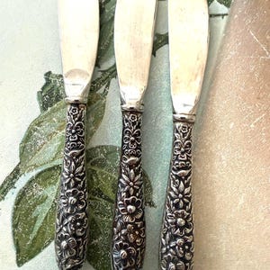 May include: Three silver-toned butter knives featuring smooth, rounded blades and ornate, embossed floral handles. The handles display a detailed, raised pattern of various flowers and leaves covering the entire grip area, contrasting with the plain, polished metal blades.