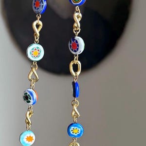 Puede incluir: Un collar de cadena dorada con cuentas de vidrio de colores. Las cuentas presentan varios patrones, incluyendo flores, estrellas y ojos. El collar tiene un cierre dorado.