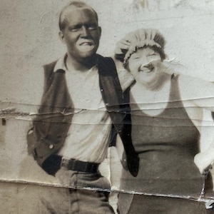 May include: Vintage black and white photograph of two people smiling. A man in a collared shirt, vest, and pants stands next to a woman in a swimsuit and swim cap. The photo is torn and aged.