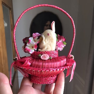 Puede incluir: Una cesta de sat&eacute;n rosa con un conejito blanco dentro. La cesta est&aacute; decorada con flores rosas y cintas.