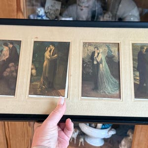 May include: A framed artwork featuring six vintage prints depicting romantic scenes. The prints, in muted tones, show couples in various poses. The black frame has a cream-colored matting. The artwork is held by a person's hand.