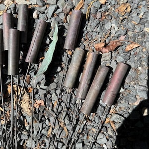 May include: A collection of metal garden stakes with cylindrical tops. The stakes are made of a dark, weathered metal and are arranged on a bed of gray gravel. Some stakes have a reddish hue.