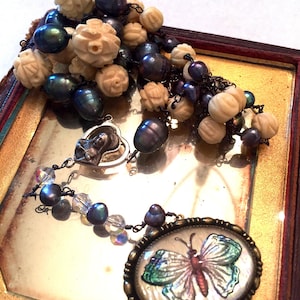 May include: An antique-style necklace with a butterfly pendant and a cluster of cream-colored carved beads and dark blue pearls. The butterfly pendant is oval-shaped with a detailed butterfly design in shades of green, red, and blue.