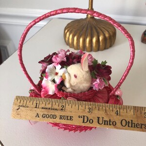 Puede incluir: Una peque&ntilde;a cesta de mimbre rosa con un conejito blanco y flores rosas y blancas en su interior. La cesta tiene un asa de sat&eacute;n rosa y una regla de madera con el texto "A Good Rule "Do Unto Others" es visible en primer plano.