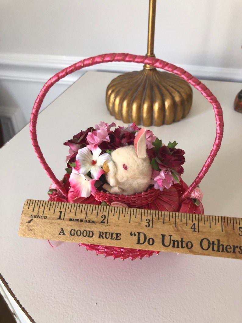 Puede incluir: Una peque&ntilde;a cesta de mimbre rosa con un conejito blanco y flores rosas y blancas en su interior. La cesta tiene un asa de sat&eacute;n rosa y una regla de madera con el texto "A Good Rule "Do Unto Others" es visible en primer plano.