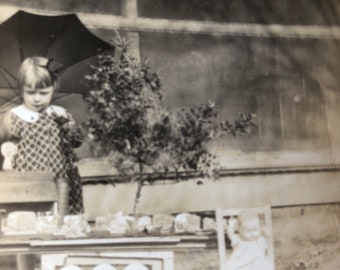 May include: A black and white photo of a young girl standing in front of a house. She is holding a small umbrella and looking at the camera. There are several framed pictures and other items on the ground in front of her.