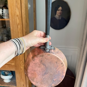 May include: A vintage copper saucepan with a long, dark handle. The pan has a rounded bottom and a tarnished, reddish-brown surface. The handle is black with a white tip and a metal loop for hanging.