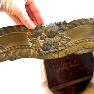 May include: An elongated, curved brass furniture mount featuring a relief design of fruit, leaves, and a textured rope-like border. The metal has a tarnished, aged patina with visible oxidation and intricate detailing across the entire surface.