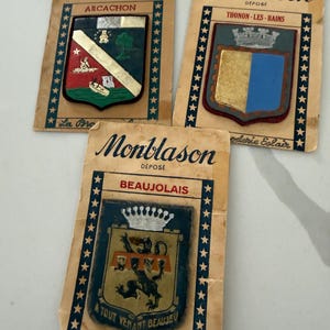 May include: Three vintage Montblason souvenir patches. Each patch features a town crest, including Arcachon, Thonon-les-Bains, and Beaujolais. The patches are on aged paper cards with star borders and the text "La Broderie Eclair".
