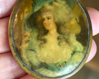 May include: Oval-shaped antique brooch featuring a portrait of a woman in a green hat and dress. The portrait is set against a yellow and brown background, with a brass-colored metal frame. The woman has curly blonde hair.