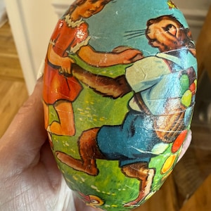 May include: Vintage egg-shaped container with a painted illustration of a girl and a rabbit in a garden setting. The egg has a light blue top and a green base with colorful details. The figures are dressed in vintage clothing.