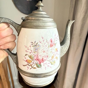May include: White ceramic teapot with a floral design featuring pink roses and other flowers. The teapot has a metal lid, handle, and spout. The handle and spout have ornate detailing.