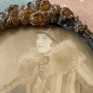 May include: Antique oval picture frame with a sepia-toned portrait of a person wearing a fur-collared coat and hat. The frame has a decorative floral design around the edge.