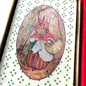 May include: A rectangular card featuring a central oval illustration of a rabbit wearing a red hood and cape, carrying a basket and a green umbrella. The card has a white background with a repeating pattern of small green dots and a gold border.
