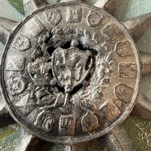 May include: A detailed, silver-toned metal starburst pendant. The pendant features a central shield design surrounded by embossed symbols and a sunburst pattern. The metal has a weathered appearance, suggesting an antique or vintage style.