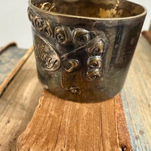 Puede incluir: Una taza de metal vintage con el nombre "Mary" inscrito en el lateral, rodeada de adornos florales. La taza tiene un aspecto deslustrado y envejecido, y se encuentra sobre una superficie desgastada y texturizada, posiblemente un libro.