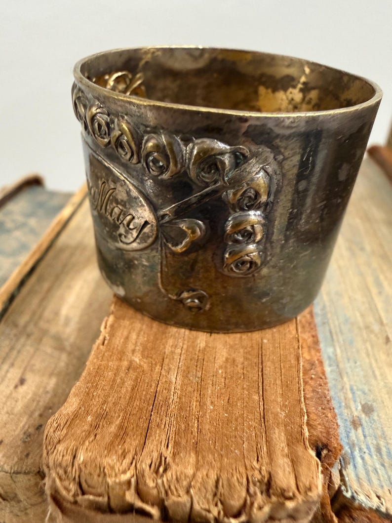 Puede incluir: Una taza de metal vintage con el nombre "Mary" inscrito en el lateral, rodeada de adornos florales. La taza tiene un aspecto deslustrado y envejecido, y se encuentra sobre una superficie desgastada y texturizada, posiblemente un libro.
