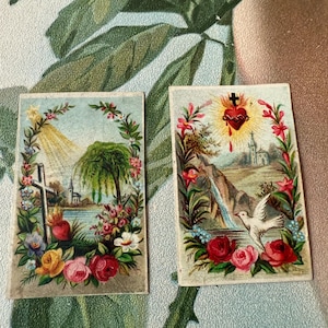 May include: Two vintage religious cards with floral borders. The first card depicts a cross, a church, and a palm tree in a landscape with a lake. The second card shows a bleeding heart with a cross, a dove, and a waterfall in a landscape with a church.