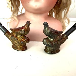 May include: Two antique bird-shaped whistles. The whistles are made of metal and feature a bird perched on a branch. The birds are painted in shades of brown, red, and gold. The whistles are black.