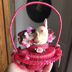 Puede incluir: Una peque&ntilde;a cesta tejida de color rosa con asa, llena de flores rosas y blancas y un conejito de fieltro blanco. La cesta est&aacute; decorada con una cinta de sat&eacute;n rosa y una rosa rosa.
