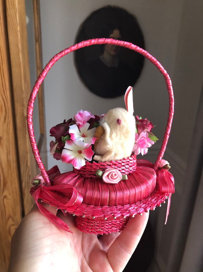Puede incluir: Una peque&ntilde;a cesta tejida de color rosa con asa, llena de flores rosas y blancas y un conejito de fieltro blanco. La cesta est&aacute; decorada con una cinta de sat&eacute;n rosa y una rosa rosa.