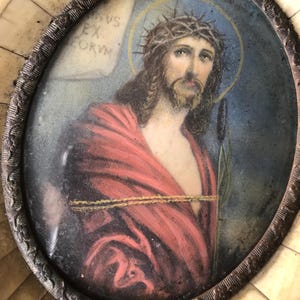 May include: An oval-shaped ivory frame with a painted image of Jesus Christ wearing a crown of thorns. The image is set against a blue background with a gold rope and a green plant.