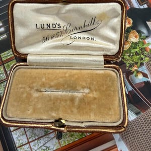 May include: Open antique jewelry box with a brown leather exterior and gold trim. The interior is lined with cream-colored satin and tan velvet. The text "LUND'S 56 & 57 Cornhill LONDON" is printed on the satin lining.