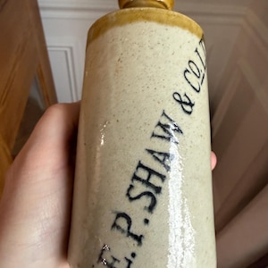 May include: A cylindrical stoneware bottle with a rounded top and a separate, rounded stopper. The bottle is beige with a darker beige rim and stopper. The text "E.P. SHAW & CO" is printed in black on the side.