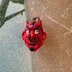 May include: A small, red devil head pendant with black accents. The pendant features horns, a mustache, and a goatee. The eyes are white. A gold-colored loop is attached to the top for hanging. The background is a textured, neutral color.