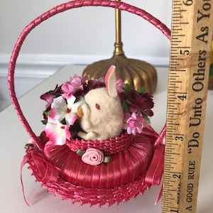 Puede incluir: Una cesta de sat&eacute;n rosa con un conejito blanco y flores rosas y burdeos en su interior. La cesta tiene un asa de sat&eacute;n rosa y un ribete dorado.